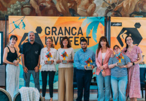 Ocho personas posan alineadas en un evento frente a un fondo con el texto “GRANCA LIVE FEST”, sosteniendo varios trofeos coloridos con formas geométricas. La escena se desarrolla en un espacio interior decorado con un mural artístico en la parte superior y paneles gráficos en tonos naranja y verde. Las personas están vestidas con ropa variada y se sitúan en fila mirando hacia la cámara, con una mesa y sillas parcialmente visibles en la parte inferior.