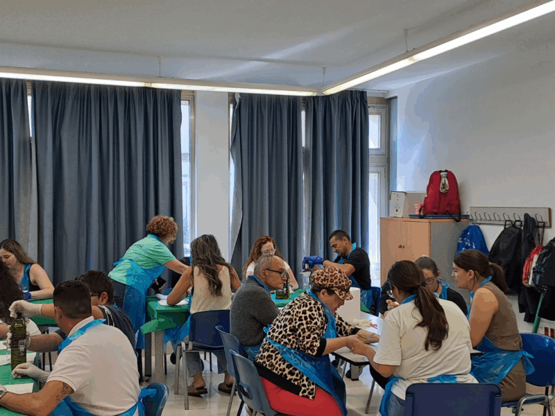 Grupo de personas participa en una actividad de voluntariado en una sala amplia con mesas cubiertas con manteles verdes. Las personas están sentadas trabajando en tareas manuales, llevando delantales azules y utilizando materiales como botellas de vidrio, utensilios y recipientes sobre las mesas. En el lado derecho se ve un roll‑up con el logotipo de Fundación Adecco y el texto “Hoy tienes la oportunidad de cambiar la vida de una persona. Atrévete”. El espacio cuenta con ventanas con cortinas grises, iluminación fluorescente y percheros con mochilas y abrigos al fondo.
