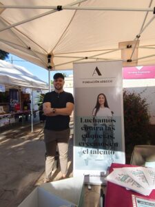 Persona de pie bajo una carpa en un evento al aire libre, situada junto a un roll‑up de Fundación Adecco que incluye una imagen y el texto “Luchamos contra las etiquetas, creemos en el talento”. En el entorno se ven otros puestos, mesas con material informativo y carteles, así como un jardín con plantas al fondo.