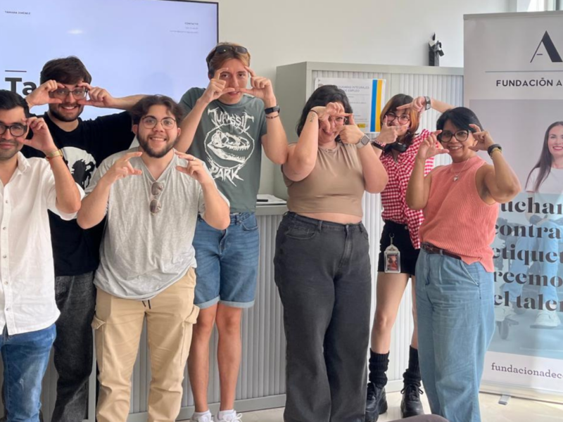 Los participantes posando en un espacio interior luminoso junto a un roll‑up de Fundación Adecco con el mensaje “Luchamos contra las etiquetas, creemos en el talento”. Las personas están de pie frente a una pantalla y realizan un gesto con las manos formando un marco alrededor de los ojos. La sala cuenta con grandes ventanales que dejan entrar luz natural, mobiliario de oficina y elementos de formación o reunión en el fondo.