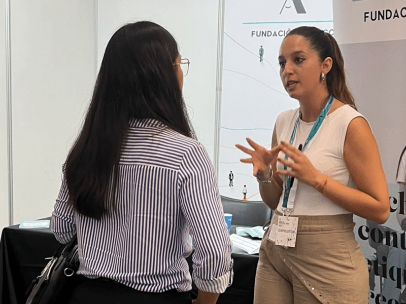 Mireia informando a otra persona en un estand de Fundación Adecco durante la feria. En el espacio se observan elementos corporativos de la fundación y material promocional con el mensaje: “Luchamos contra las etiquetas, creemos en el talento”. Ambas personas conversan de pie frente al expositor.