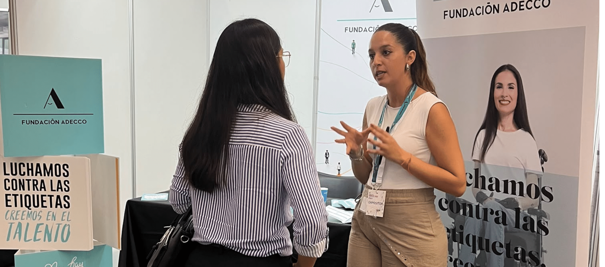 Mireia informando a otra persona en un estand de Fundación Adecco durante la feria. En el espacio se observan elementos corporativos de la fundación y material promocional con el mensaje: “Luchamos contra las etiquetas, creemos en el talento”. Ambas personas conversan de pie frente al expositor.